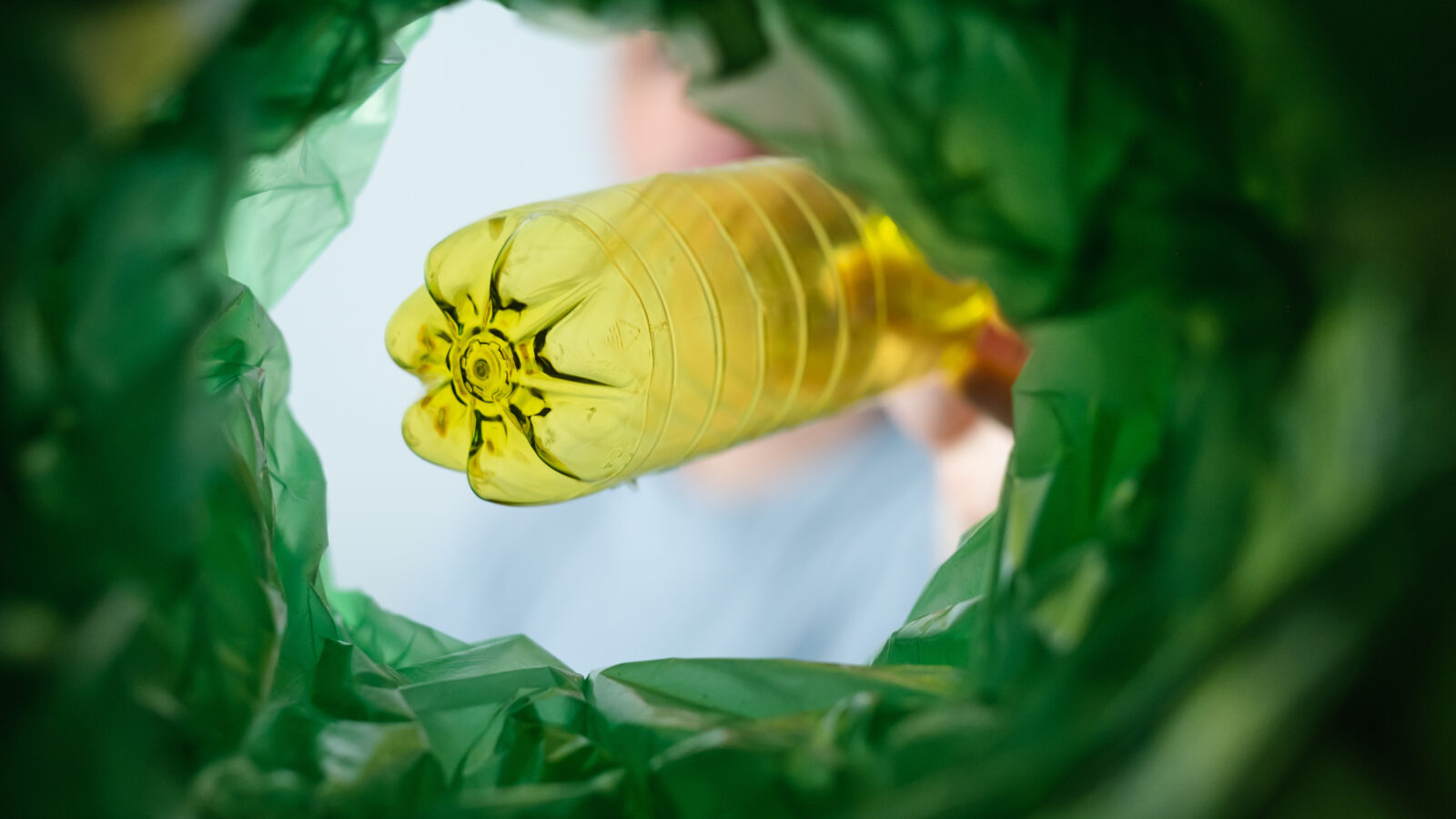 A yellow plastic drinks bottle is being lowered into a recycling bin. The picture is taken from inside the green plastic refuse sack, looking up towards the plastic bottle.