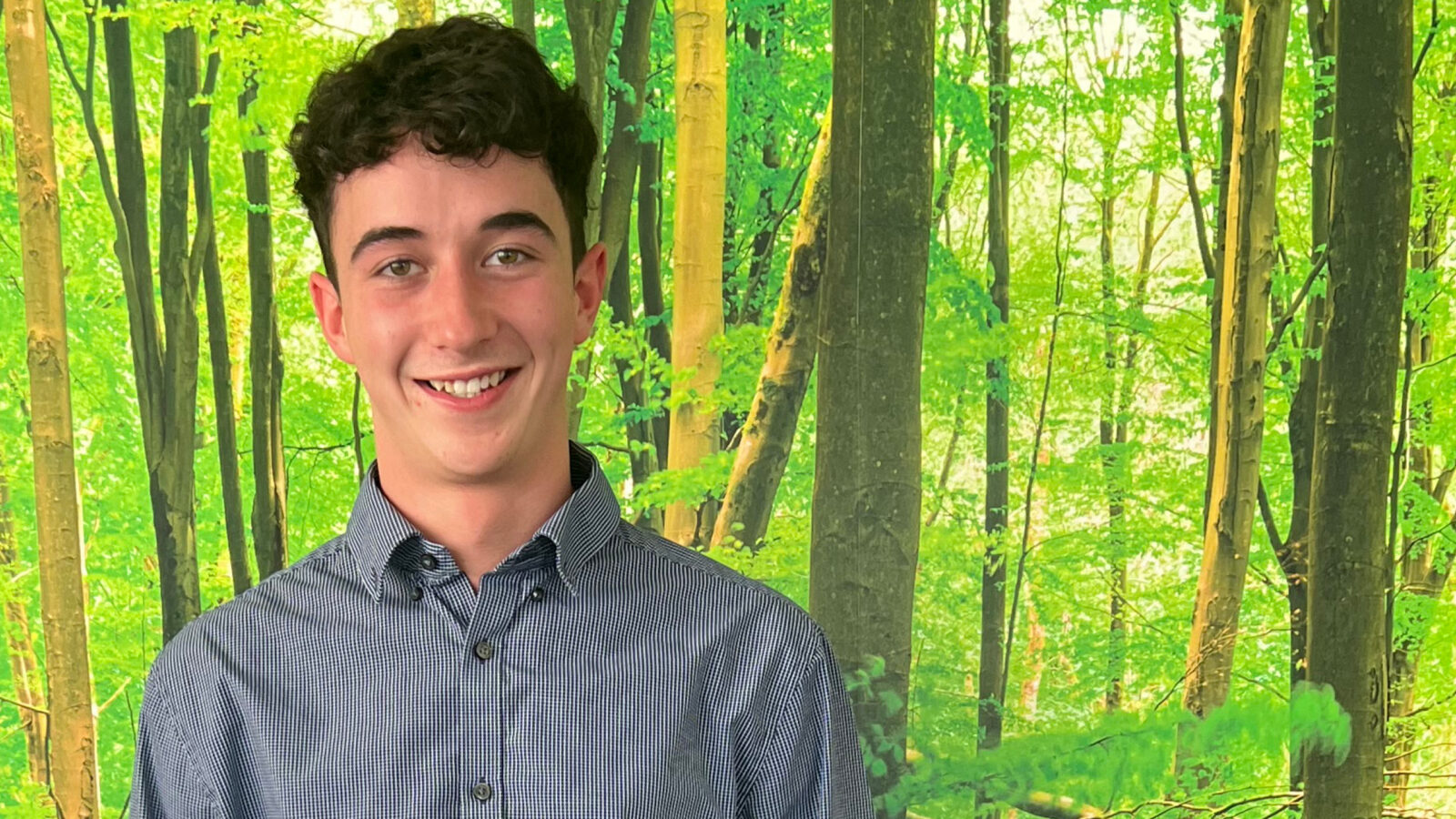 Endurance Vehicle Solutions apprentice, Archie is pictured standing against a photo wallpaper of a bright and vibrant woodland.