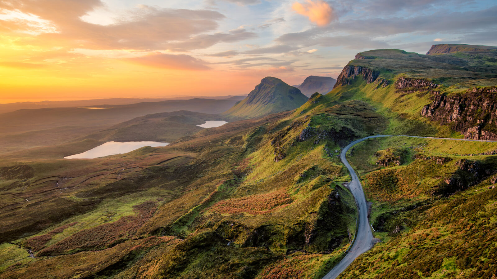 Rolling Scottish hillside, against an orange sunset. A road climbs up the nearest hill.